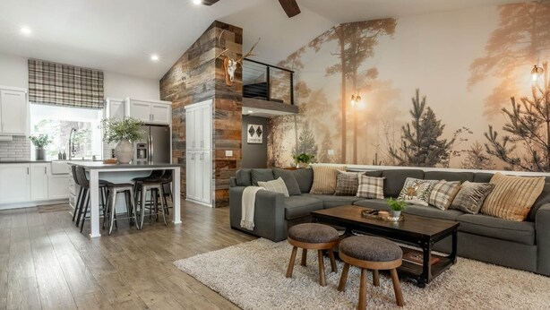 Ein modernes Wohnzimmer mit grauem Sofa, einem Holztisch und einer Küchenzeile mit weißen Schränken und einer Wand in Steinoptik.