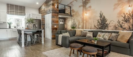 Un salon moderne avec un canapé gris, une table basse en bois et un coin cuisine doté de meubles blancs et d’un mur d’accent en pierre.