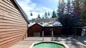 Eine Holzterrasse mit Whirlpool, Metallgeländer und Blick auf ein Gebäude mit geneigtem Dach und Schnee.