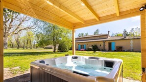 A wooden deck with a hot tub, a covered area, and a stone building with blue shutters in the background. - Domaine des Bruyères | Workshop (Saint-Savin)
