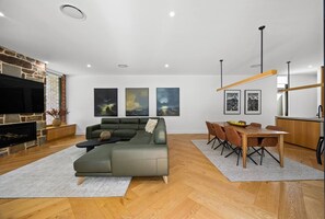 Un salon moderne avec un canapé d’angle, un coin repas avec une table et des chaises en bois, et une cuisine dotée d’un luminaire suspendu.
