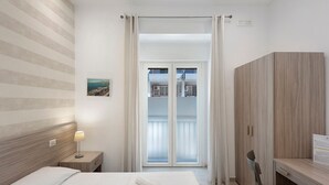 Quarto de hotel com cama, escrivaninha, cadeira, mesa de cabeceira, luminária, janela com cortinas e vista para uma varanda.
