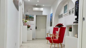 Um corredor com uma cadeira vermelha, paredes brancas e uma porta que dá para outro quarto.