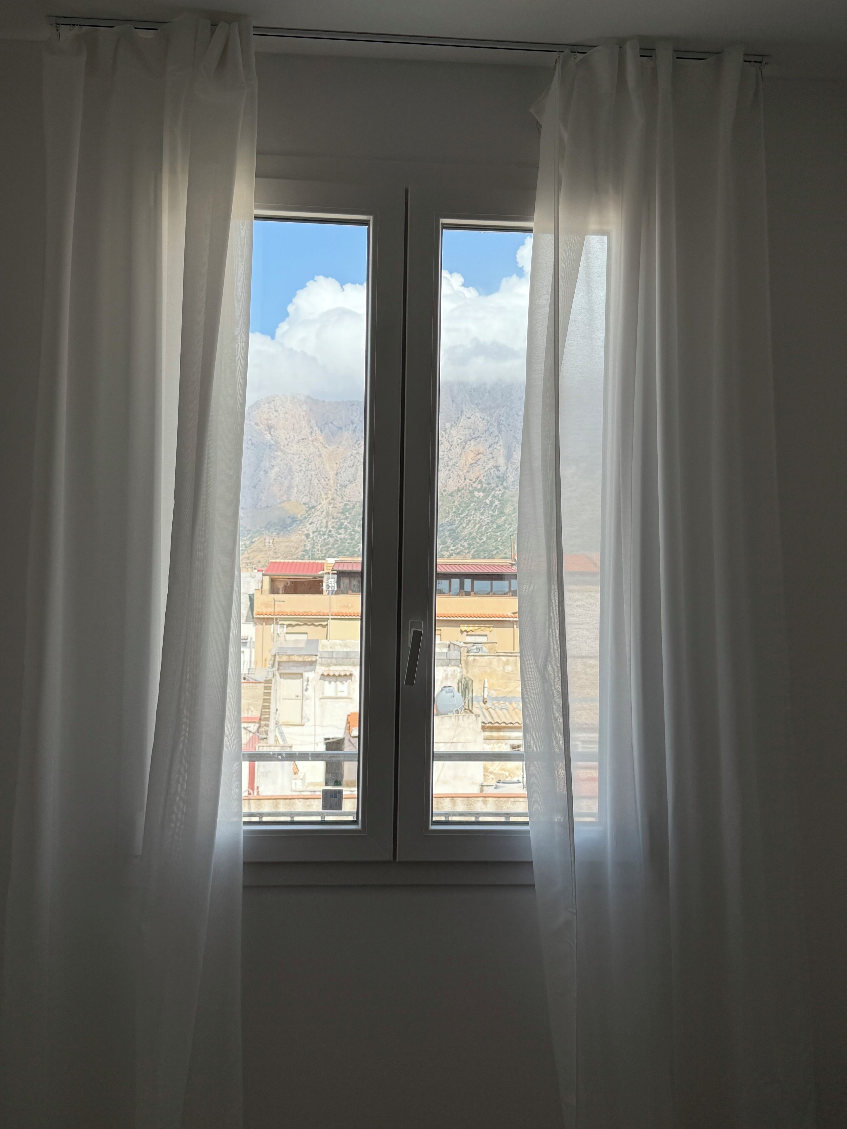 A room with a large window featuring white curtains, offering a view of a mountainous landscape and buildings below.