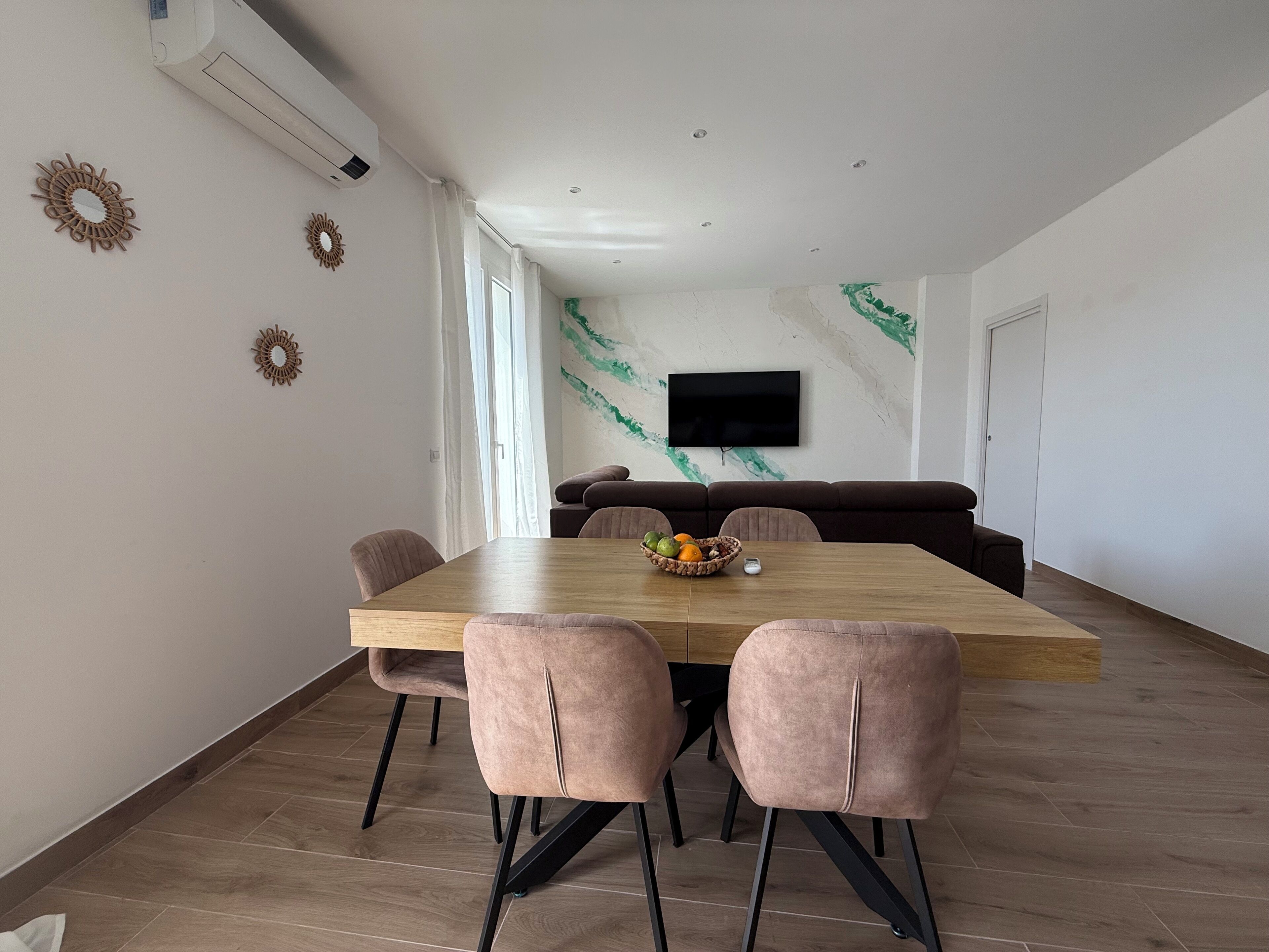 Un espace repas moderne avec une table en bois, des chaises rembourrées, un panier de fruits et un téléviseur fixé au mur.