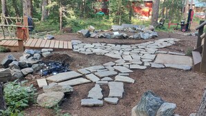 Kivipolku johtaa metsäisen alueen läpi, ja taustalla näkyy puinen rakennelma sekä traktori.