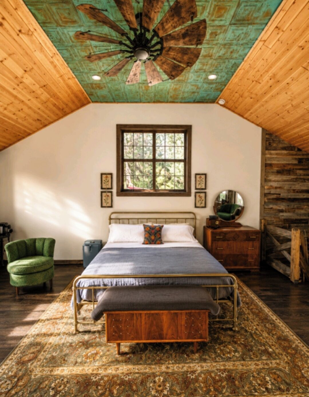 A bedroom with a bed, a wooden ceiling fan, a window, a green chair, a bedside table, and a mirror.