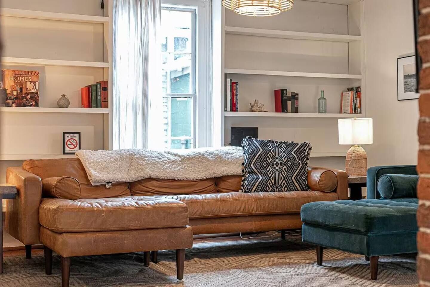 Un salon avec un canapé en cuir marron, un fauteuil bleu et un coussin décoratif à motifs. On y trouve des étagères remplies de livres et d’objets décoratifs, ainsi qu’une lampe sur une table d’appoint.