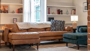 Una sala de estar con un sofá de cuero marrón, un sillón azul y un cojín estampado. Se ven estanterías con libros y objetos decorativos, y una lámpara sobre una mesita auxiliar.