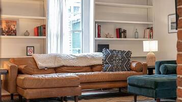 Un salon avec un canapé en cuir marron, un fauteuil bleu et un coussin décoratif à motifs. On y trouve des étagères remplies de livres et d’objets décoratifs, ainsi qu’une lampe sur une table d’appoint.