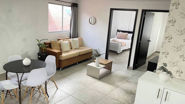 Een moderne woonkamer met een bank, eettafel en stoelen. Er staat een klein bijzettafeltje met een vaas. Een spiegel hangt aan de muur, en een deur leidt naar een andere kamer.