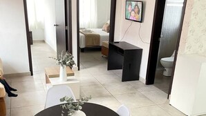 Een eethoek met een ronde tafel, witte stoelen en een vaas met bloemen. Een televisie hangt aan de muur en er is een bed te zien door een open deur.