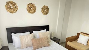 Een net opgemaakt bed met een geborduurde witte deken, meerdere kussens en een bruin kussen met structuur. Boven het bed hangen drie decoratieve objecten. Een bruine bank met een wit kussen staat tegen de muur, en een klein tafeltje met een lamp staat naast het bed.