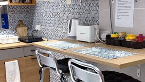 Une cuisine avec un dosseret carrelé, une machine à café, un grille-pain et un petit réfrigérateur. On y trouve deux tabourets de bar et une serviette blanche accrochée à la porte.