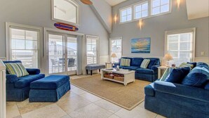 A spacious living room with a high ceiling, large windows, and a view of the outdoors. - Aloha from Kure South (Kure Beach)
