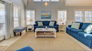 A spacious living room with a high ceiling, large windows, and a cozy blue sofa set. - Aloha from Kure South (Kure Beach)