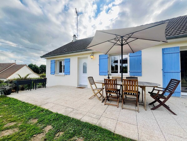 A house with a patio, a dining table with chairs, and a large umbrella. - Le Logis des Pres, Country Stay (Bricquebosq)