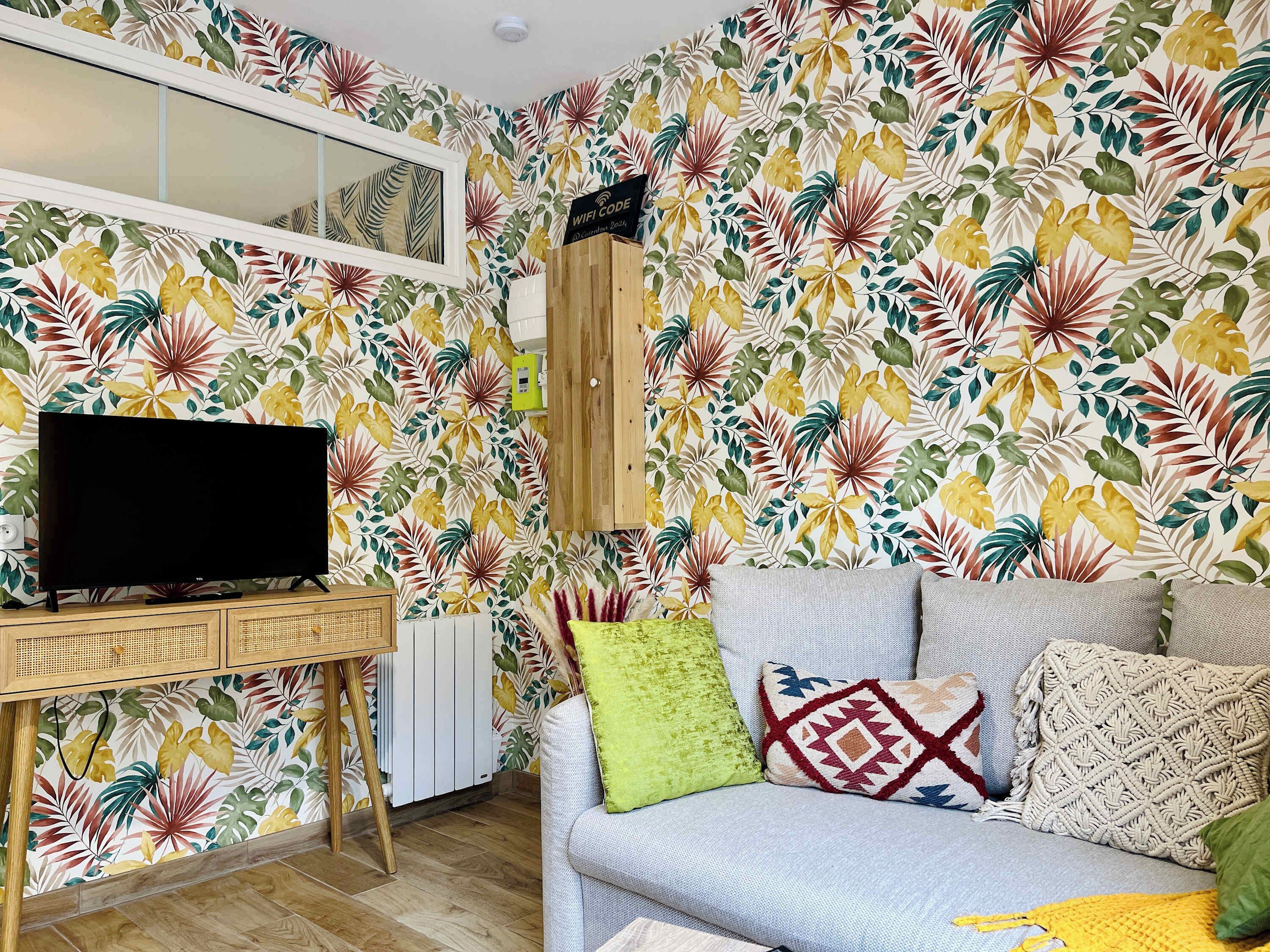 Een woonkamer met een flatscreen televisie, een houten tv-meubel, een bank met opvallende kussens en een muur met tropische bladbehang.