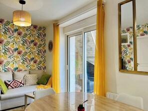 Une pièce avec un mur orné de motifs, une table ronde, un canapé avec des coussins, un luminaire suspendu et une porte-fenêtre coulissante.