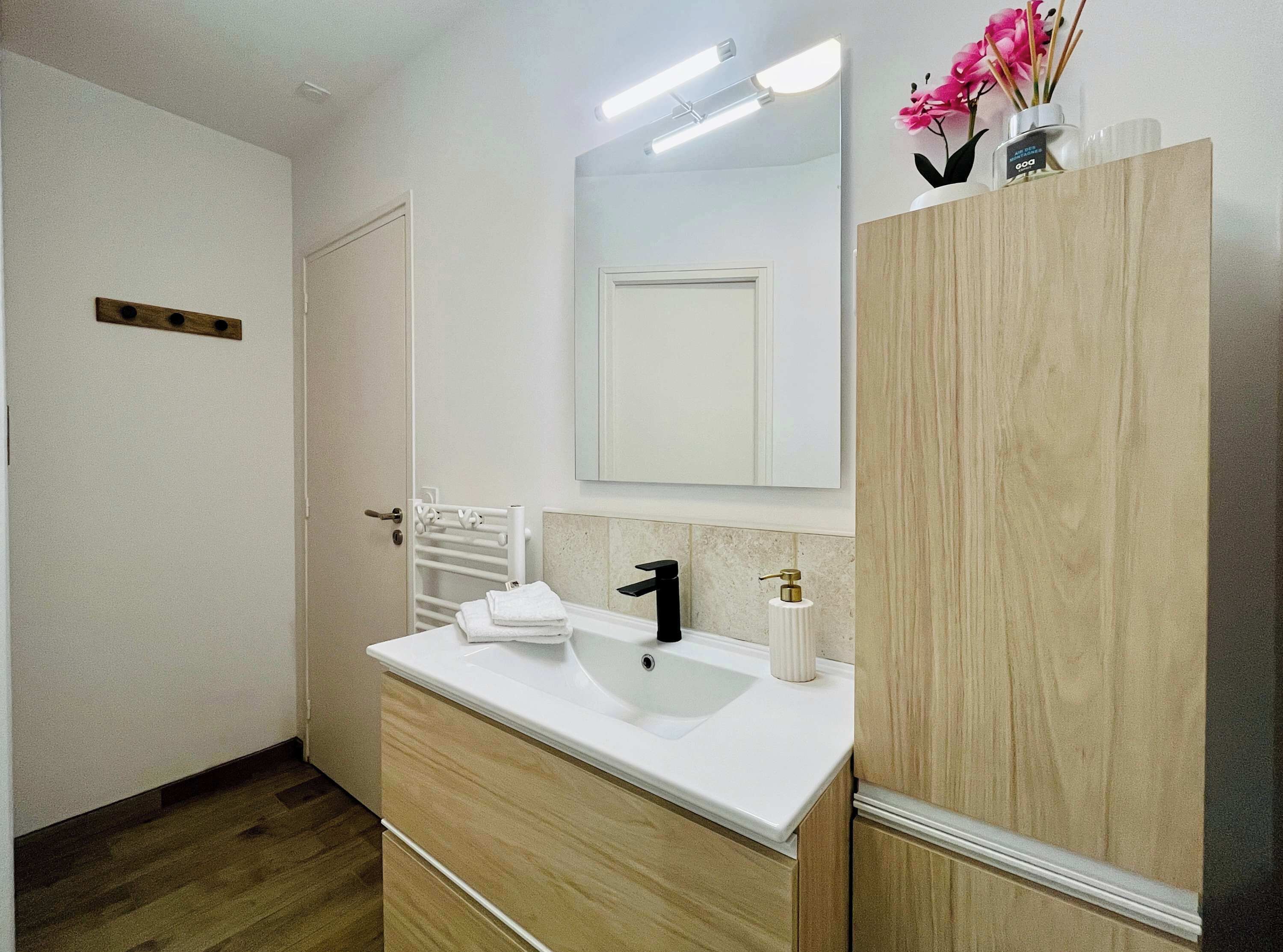 Un baño con lavamanos blanco, grifo negro y espejo arriba. Hay un perchero y una botella de jabón para manos en la encimera.