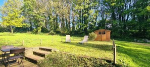 Eine Holzterrasse mit Tisch und Stühlen, ein kleines Gartenhaus und eine Rasenfläche mit Bäumen.