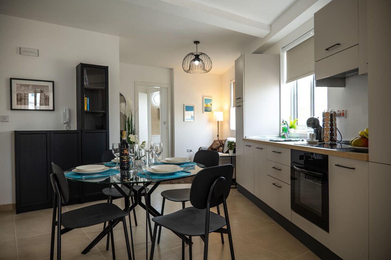A modern kitchen with a dining area, featuring a glass-top table, black chairs, and a pendant light.
