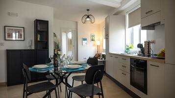 A modern kitchen with a dining area, featuring a glass-top table, black chairs, and a pendant light.