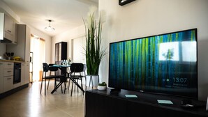 A modern living room with a large flat-screen TV on a black stand, a dining area with a table and chairs, and a kitchen in the background. - Rosie - Two Bedroom Apartment (Pietà)
