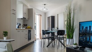 A modern kitchen with white cabinets, a dining area with a table and chairs, a large TV on a stand, and a decorative plant. - Rosie - Two Bedroom Apartment (Pietà)