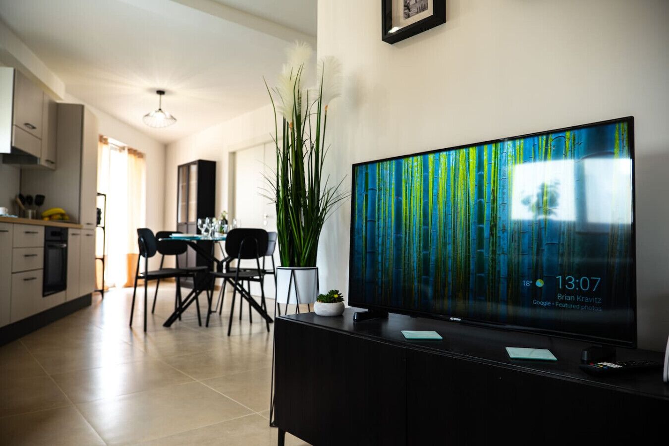 Un soggiorno moderno con una grande TV a schermo piatto su un supporto nero, una zona pranzo con tavolo e sedie, e una cucina sullo sfondo.