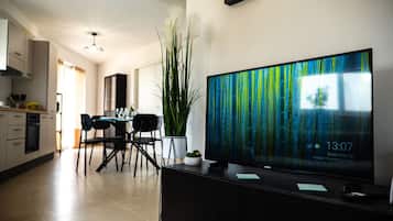 Un soggiorno moderno con una grande TV a schermo piatto su un supporto nero, una zona pranzo con tavolo e sedie, e una cucina sullo sfondo.