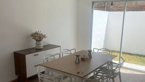 Een eetruimte met een houten tafel en witte stoelen, een klein kastje met een vaas gedroogde bloemen, en een groot raam met uitzicht op een bakstenen muur.
