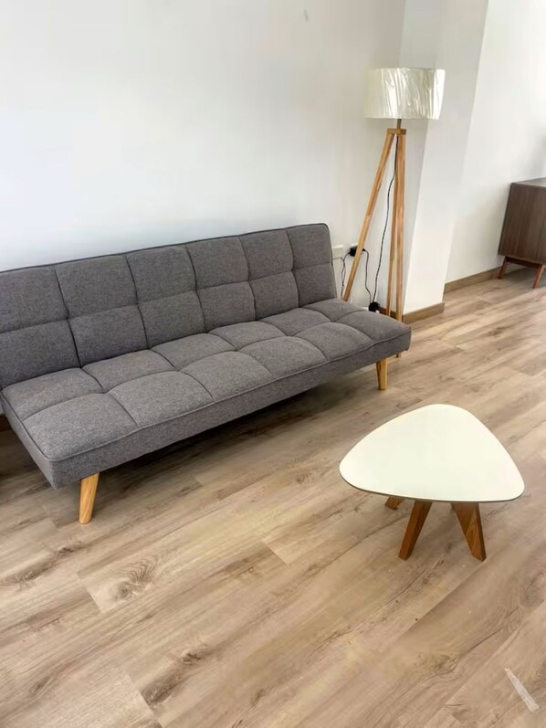 Un canapé gris aux pieds en bois et une table blanche aux pieds en bois, dans une pièce dont le sol est recouvert de parquet.