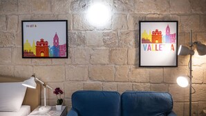 A room with a stone wall, two framed cityscape pictures, a white lamp, a blue sofa, and a small potted plant. - Casa San Carlo Valletta - Amarillo (Valletta)
