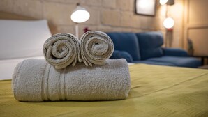 A neatly made bed with a rolled towel placed on top, a blue sofa in the background, and a lamp on the nightstand. - Casa San Carlo Valletta - Amarillo (Valletta)