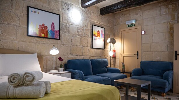 A hotel room with a bed, two blue sofas, a small table, and two wall-mounted lamps. - Casa San Carlo Valletta - Amarillo (Valletta)