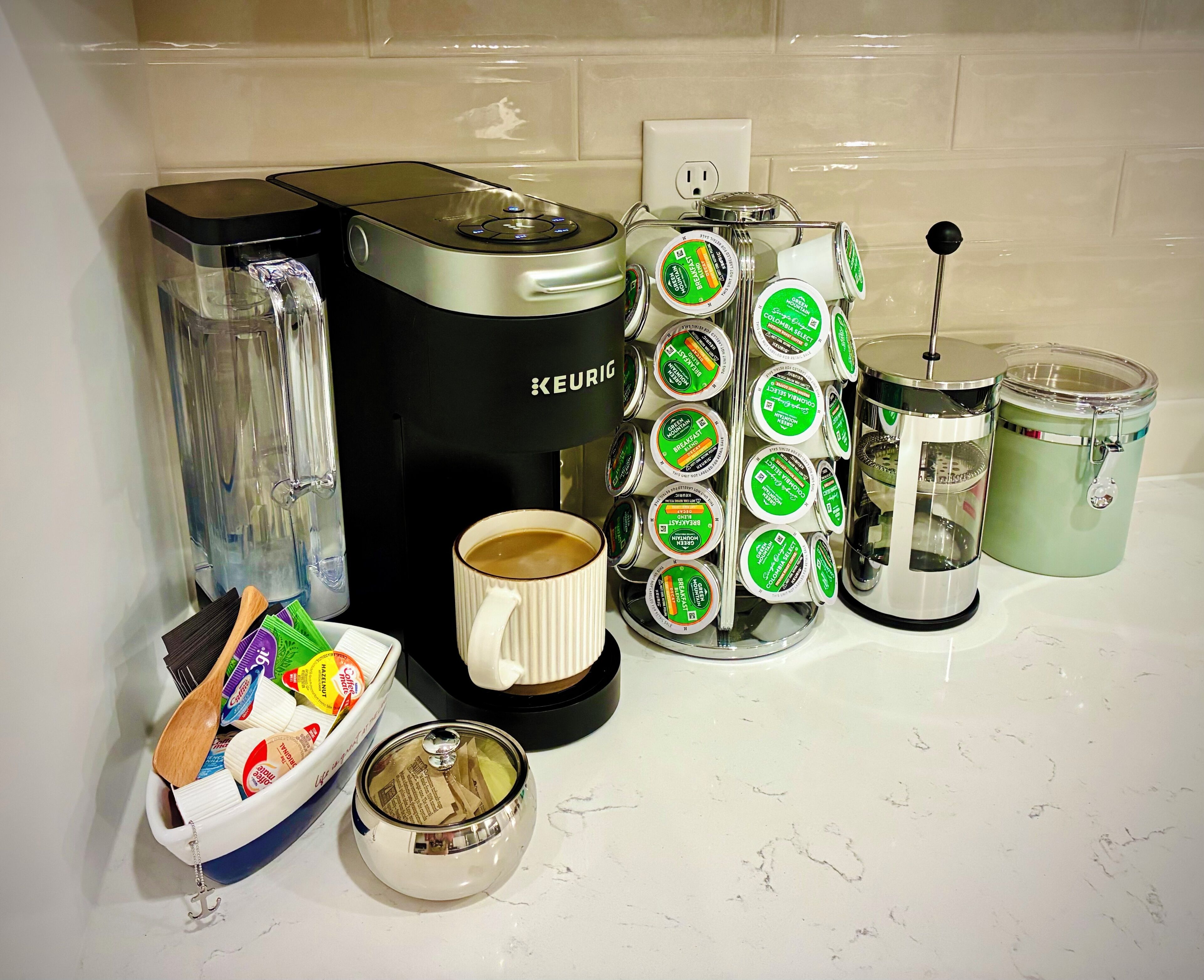 Una macchina per caffè Keurig, una tazza da caffè, un macinino per caffè e un contenitore di cialde per caffè su un piano di lavoro in cucina.