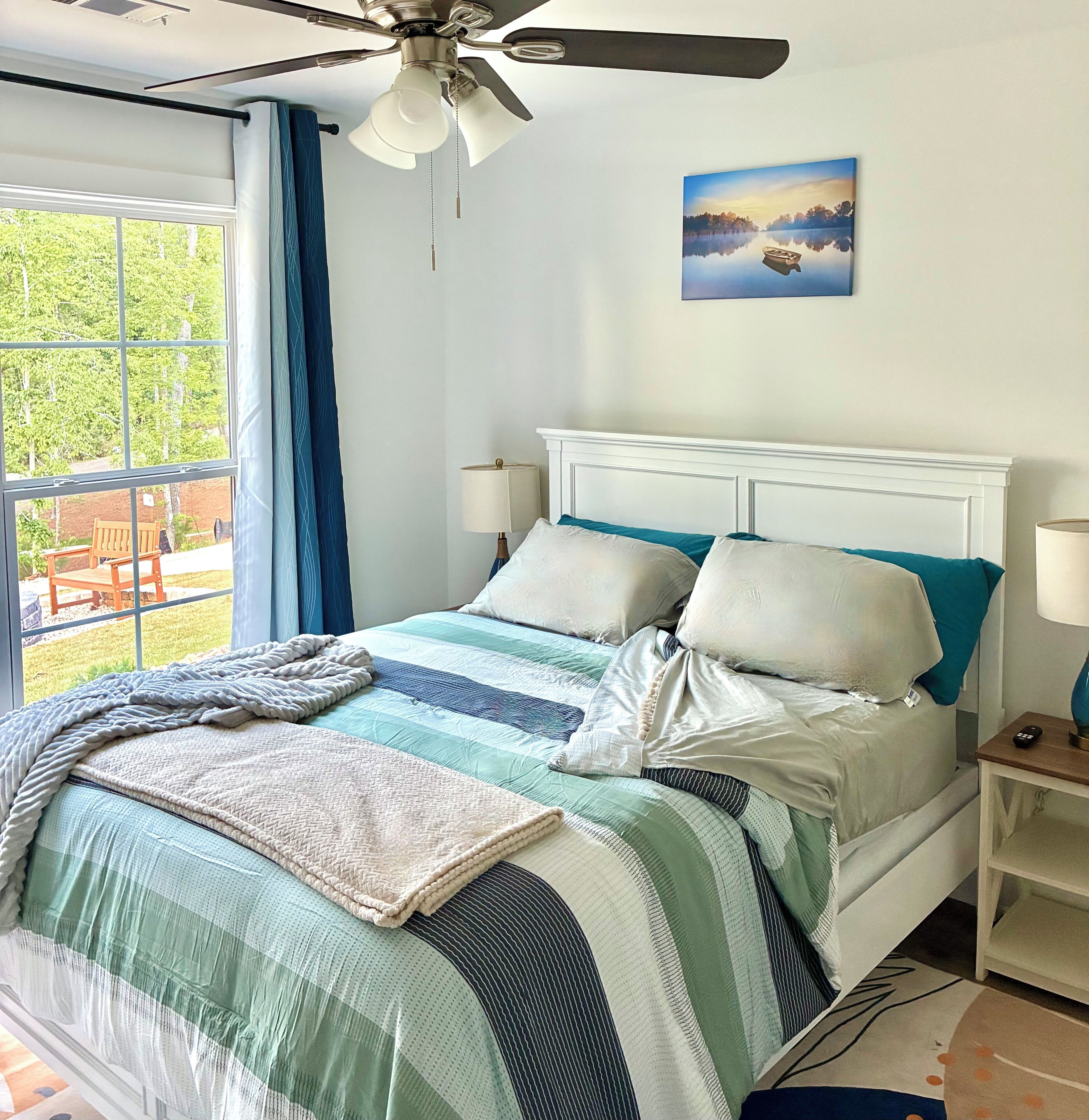 A neatly made bed with a striped bedspread, a ceiling fan, a nightstand with a lamp, and a painting of a lake scene.