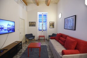 A living room with a television, a red sofa, a blue armchair, a red table, and a window with shutters. - The Lascaris Suite at Grand Harbour View Flats, (Il-Furjana)