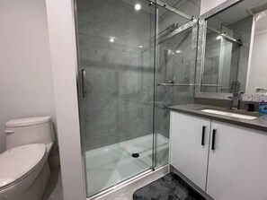 A modern bathroom with a glass-enclosed shower, a white toilet, and a sink with a mirror above it. - The Theatre Suite (Delta)