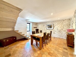 Uma área de refeições com piso de azulejos hexagonais, uma mesa de madeira e cadeiras. Uma escada fica à esquerda e uma parede de pedra à direita.