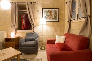A cozy room with a grey armchair, a red sofa, a wooden nightstand with a lamp, a window with curtains, and a framed cityscape painting on the wall. - Casa San Carlo Valletta - Rosa (Valletta)