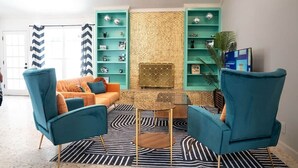 Una sala de estar con sillones color turquesa, un sofá naranja y un tapete estampado. Se observan estanterías con objetos decorativos y una chimenea.