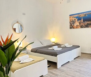 Una camera d'albergo con due letti, uno specchio rotondo, una lampada da comodino, una pianta e un quadro raffigurante un panorama urbano.