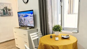 Kalustettu huone, jossa on televisio, pieni pöytä keltaisella pöytäliinalla, ruukkukasvi ja harmaa, pehmustettu tuoli.