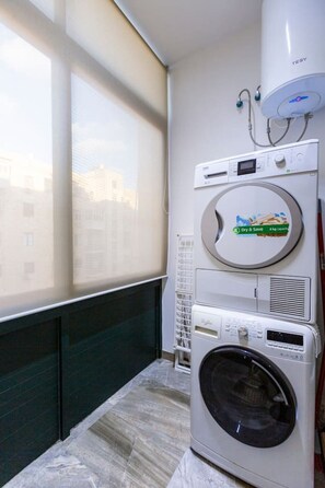 A compact laundry room with a front-loading washer and dryer, a window with blinds, and a radiator. - Valletta - Deluxe 3 Bedroom Apartment (Il-Belt Valletta)