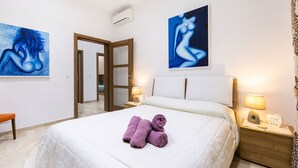Habitación de hotel con cama, dos cuadros en las paredes, un banco y una mesita de noche con lámpara.
