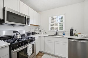Een moderne keuken met witte kastjes, roestvrijstalen apparatuur, een waterkoker op het fornuis en een raam met zonwering.
