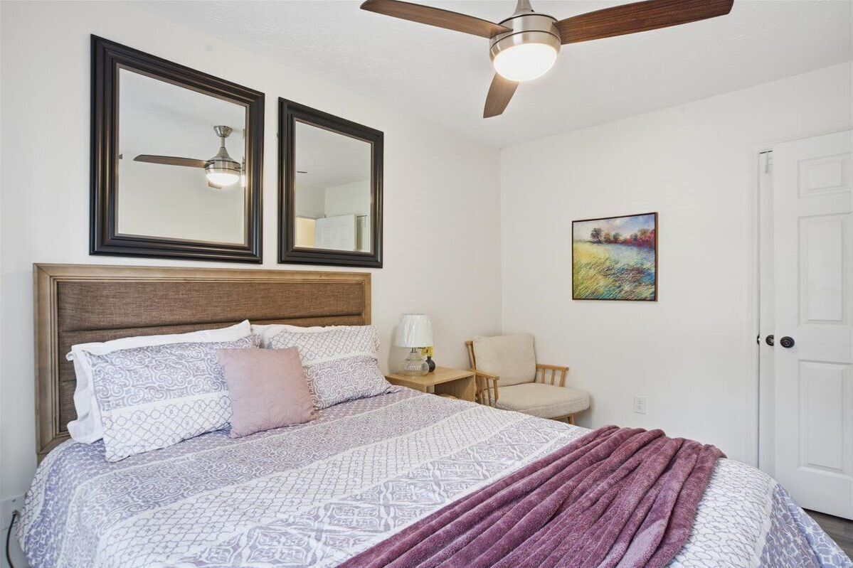 A bedroom with a bed, two mirrors, a ceiling fan, a nightstand, a chair, and a door.