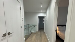 A modern hallway with wooden flooring, white walls, and a glass railing. There is a bed visible in the adjacent room.